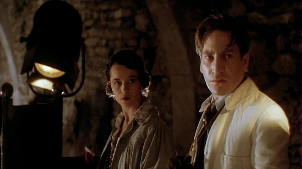 Shadow of the Vampire (2000) movie still: medium — A dark stone cellar room with rough textured walls and minimal furniture visible. A man in a…