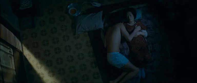 Mother (2009) movie still: wide — A dark bedroom with patterned floor tiles and a bed occupying one side. A young man lies with his arm around an…