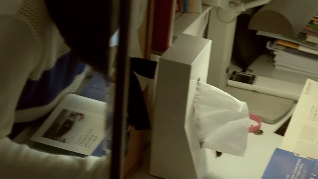 REM (2019) movie still: medium — A cluttered desk workspace holds a white tissue box and scattered papers. A person in a patterned knit sweater…