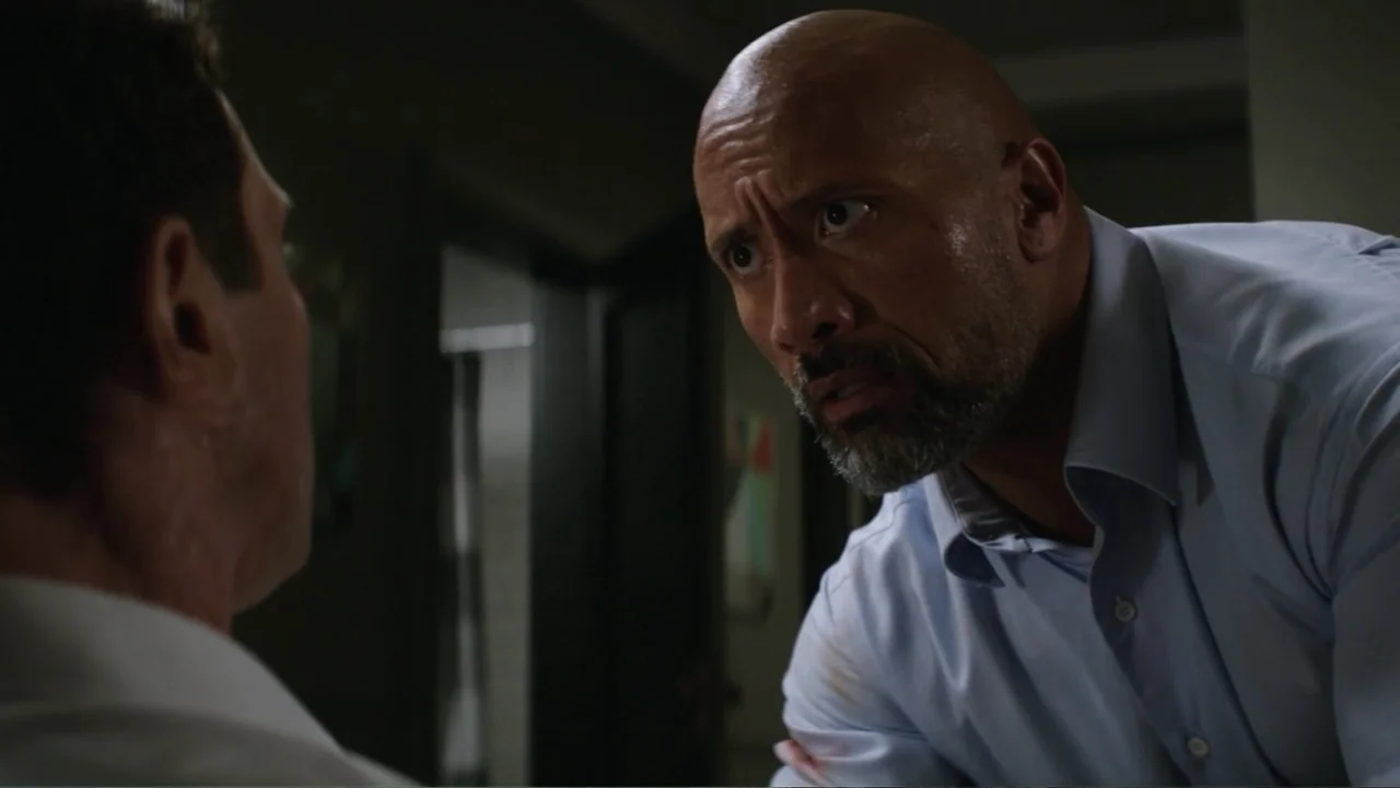Skyscraper (2018) movie still: medium — A dimly lit room with shadows obscuring the background. A man leans in closely to speak with an individual…