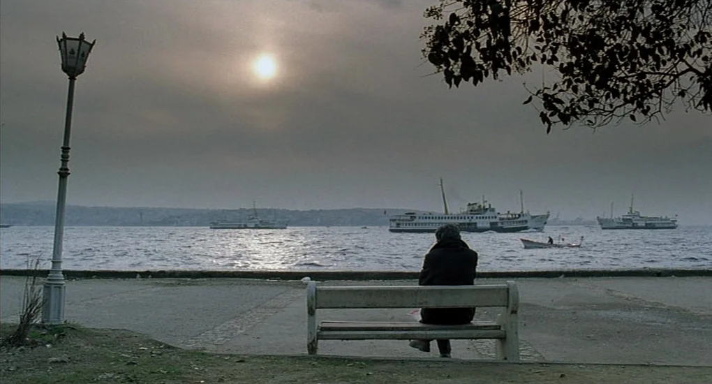 Uzak (2002) movie still: wide — A stone paved waterfront with a single bench overlooking a wide channel of water. A person sits alone on the bench…