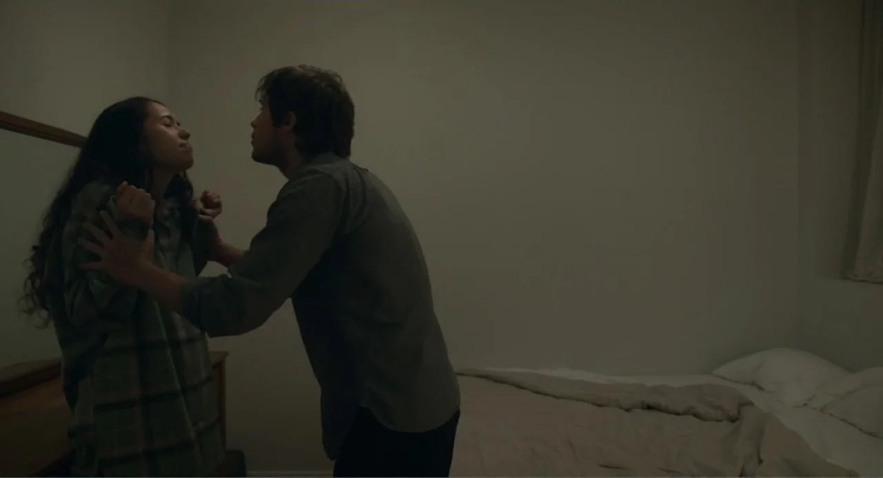 This Closeness (2024) movie still: medium — A spartan bedroom contains a bed with light-colored linens against a plain wall. A man stands…