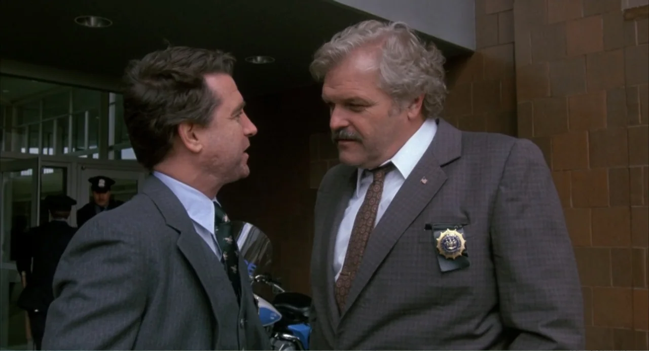 F/X (1986) movie still: medium — A building exterior entrance features brown tiled walls and a large glass door. Two men in suits stand near each…