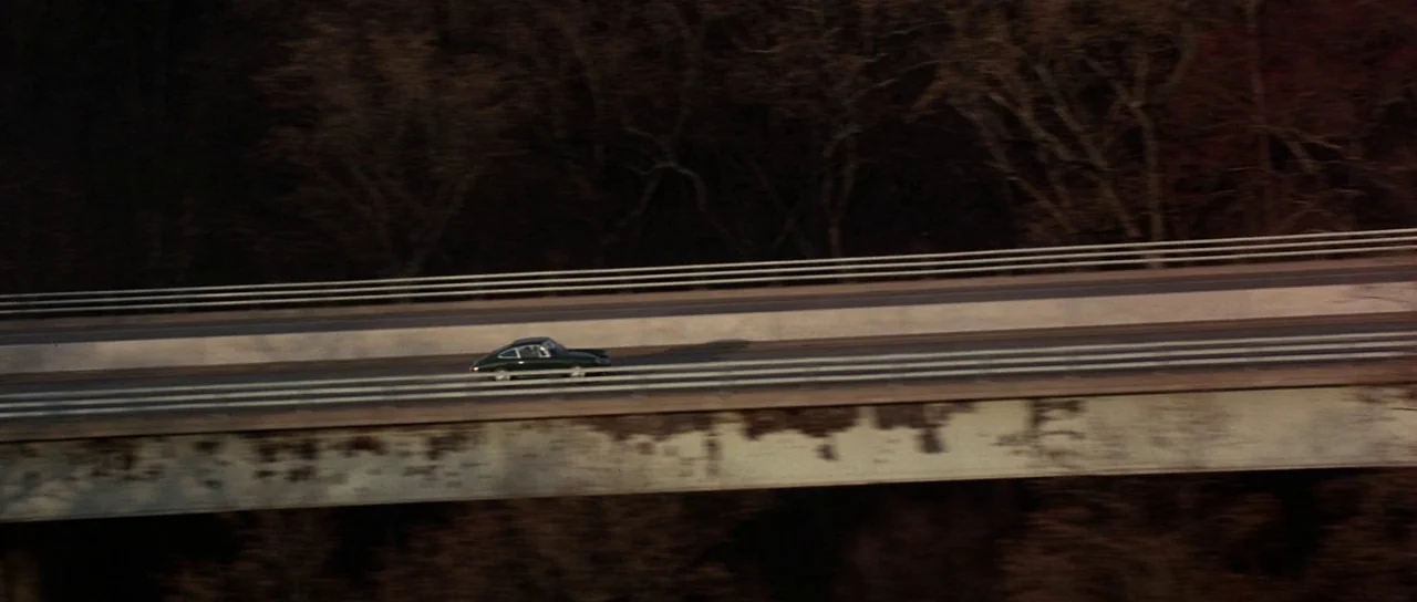 Spy Game (2001) movie still: wide — A long concrete highway bridge spans a wooded area during the late afternoon. A small dark car drives…