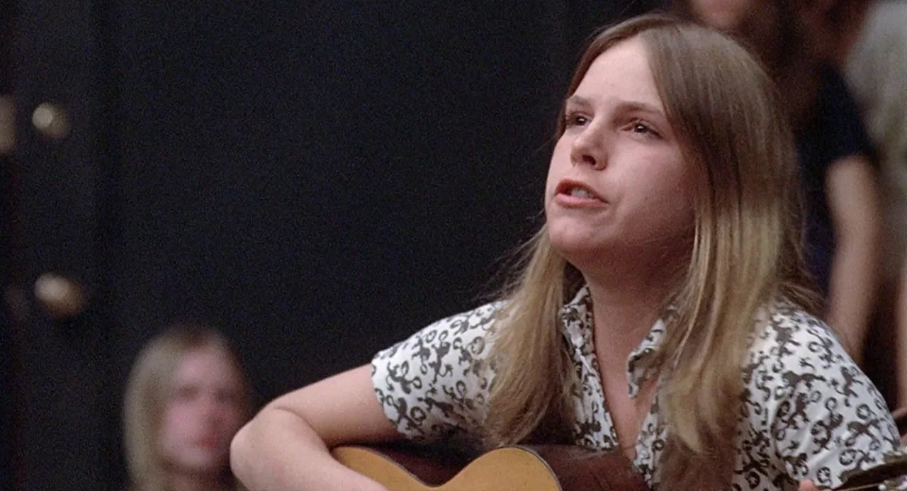 Taking Off (1971) movie still: medium — A dark indoor space with blurred figures in the background. A teenage girl with long hair plays an acoustic…