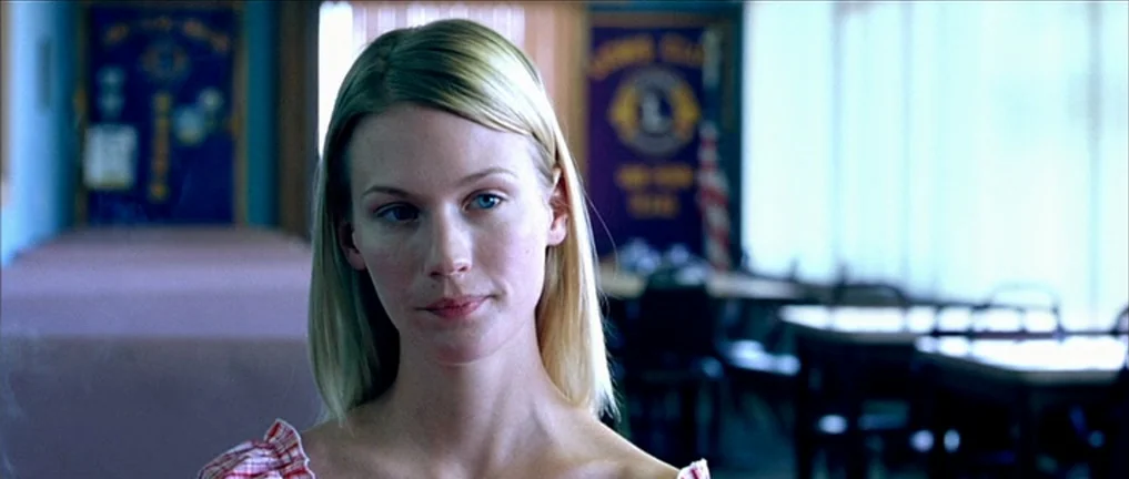 The Three Burials of Melquiades Estrada (2005) movie still: closeup — A community hall with tables and a banner visible in the blurred background. A…