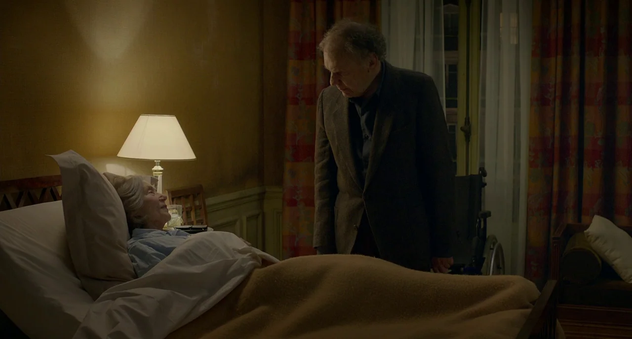 Amour (2012) movie still: medium — A dim room contains a hospital-style bed and a dark-clothed man standing beside it. The man watches a woman who…
