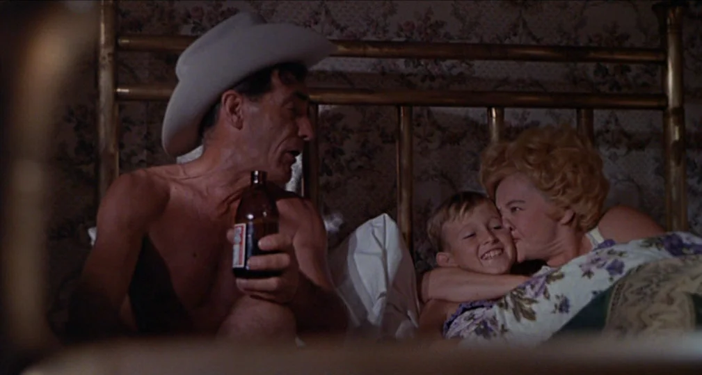 Midnight Cowboy (1969) movie still: medium — A dimly lit room features a brass bed frame with a floral wallpaper backdrop. A man in a cowboy hat…
