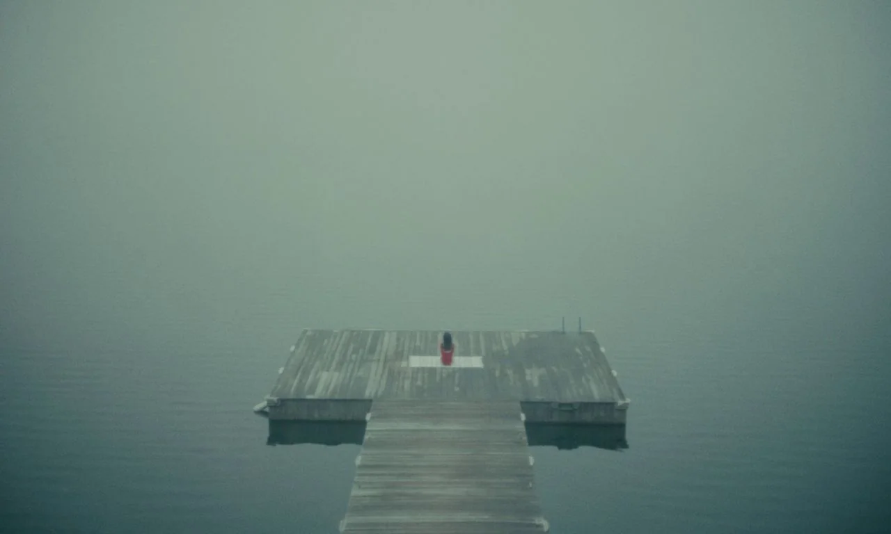 Black Bear (2020) movie still: wide — A wooden dock extends into a calm, dark lake shrouded in dense gray fog. A small figure in a red dress sits…
