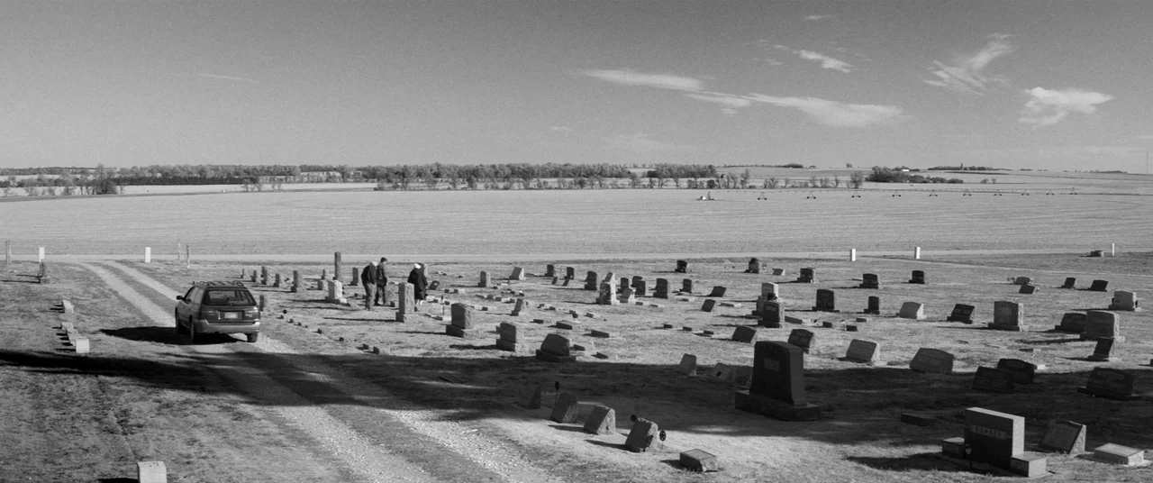 Nebraska (2013) movie still: wide — An open rural cemetery featuring many stone grave markers on a flat dry lawn. Three people walk near a parked…