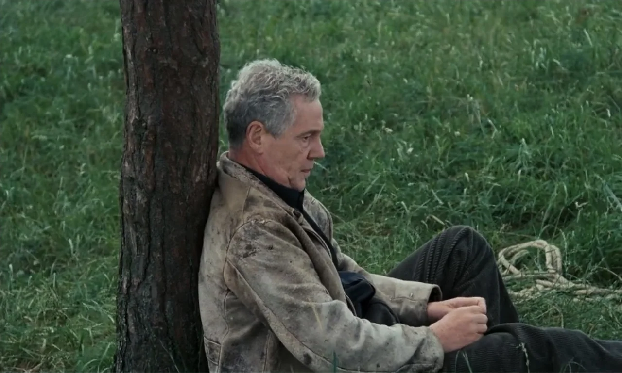 The Sacrifice (2015) movie still: medium — A man sits on a grassy field with a large tree trunk behind him. The man is wearing a dirty tan jacket…