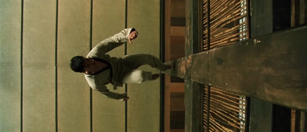 The Matrix (1999) movie still: fullbody — A traditional dojo interior features minimalist walls and a complex wooden beamed ceiling structure. A man…