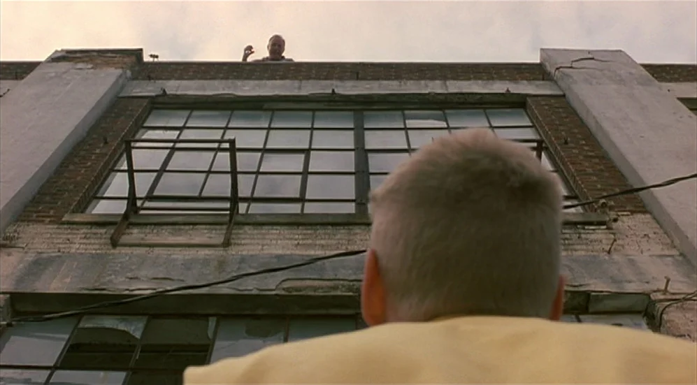 Bottle Rocket (1996) movie still: medium — A brick industrial building with multi-pane windows rises against a cloudy sky. A child in a yellow shirt…