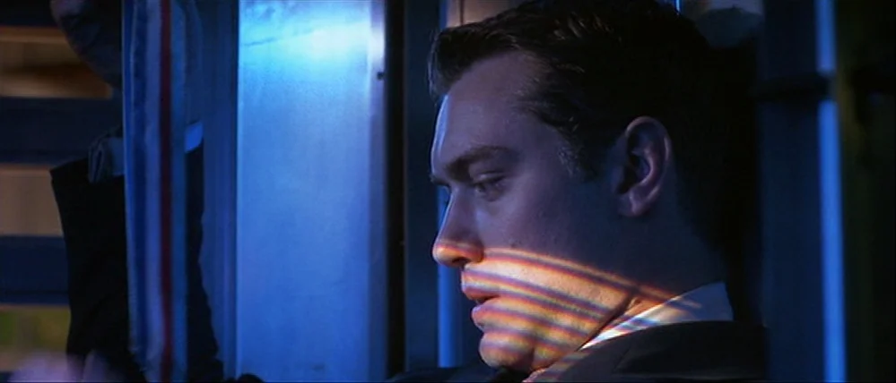 Gattaca (1997) movie still: closeup — A dark interior of a moving vehicle with blue ambient light filling the cabin. A man sits by a window as a…