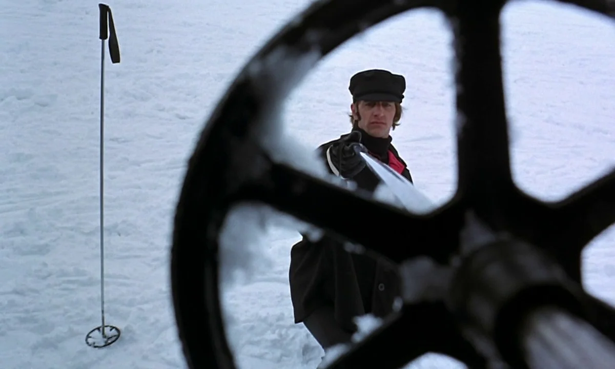 Help! (1965) movie still: medium — A man stands in an expansive, flat snowy field during the day. He holds a ski pole directly toward the camera,…