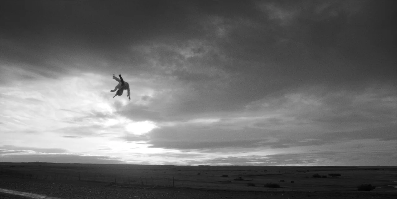 El Conde (2023) movie still: wide — A vast, desolate, and flat landscape stretches toward a cloudy horizon line. A lone figure is suspended in the…