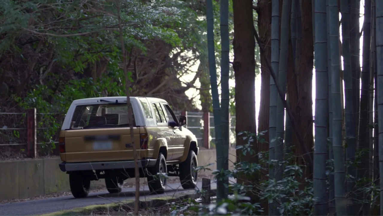 Alien Sharks: Ghosts of Japan (2024) movie still: medium — A tan SUV drives along a narrow road bordered by tall bamboo stalks. The late afternoon…
