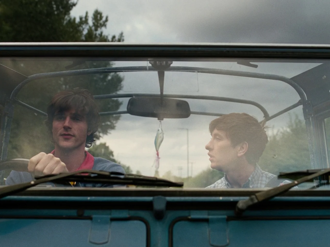 Saltburn (2023) movie still: medium — A view through the windshield of a blue, open-roof vehicle driving along a road. Two young men occupy the…