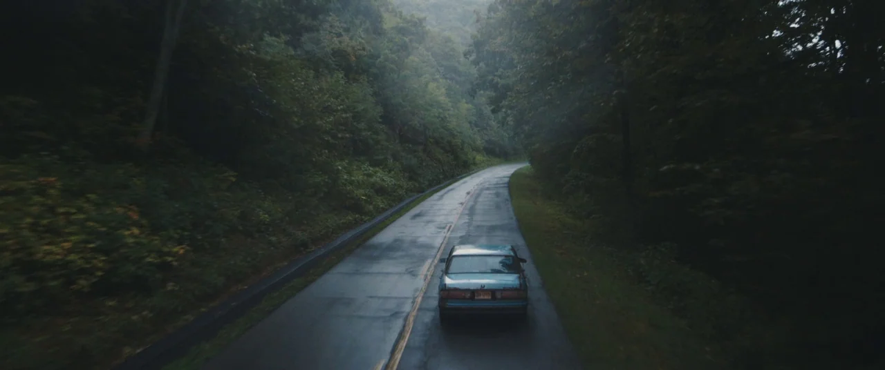 Blue Ruin (2013) movie still: wide — An asphalt road winds through a dense, wet forest under overcast gray skies. A light blue sedan drives forward…