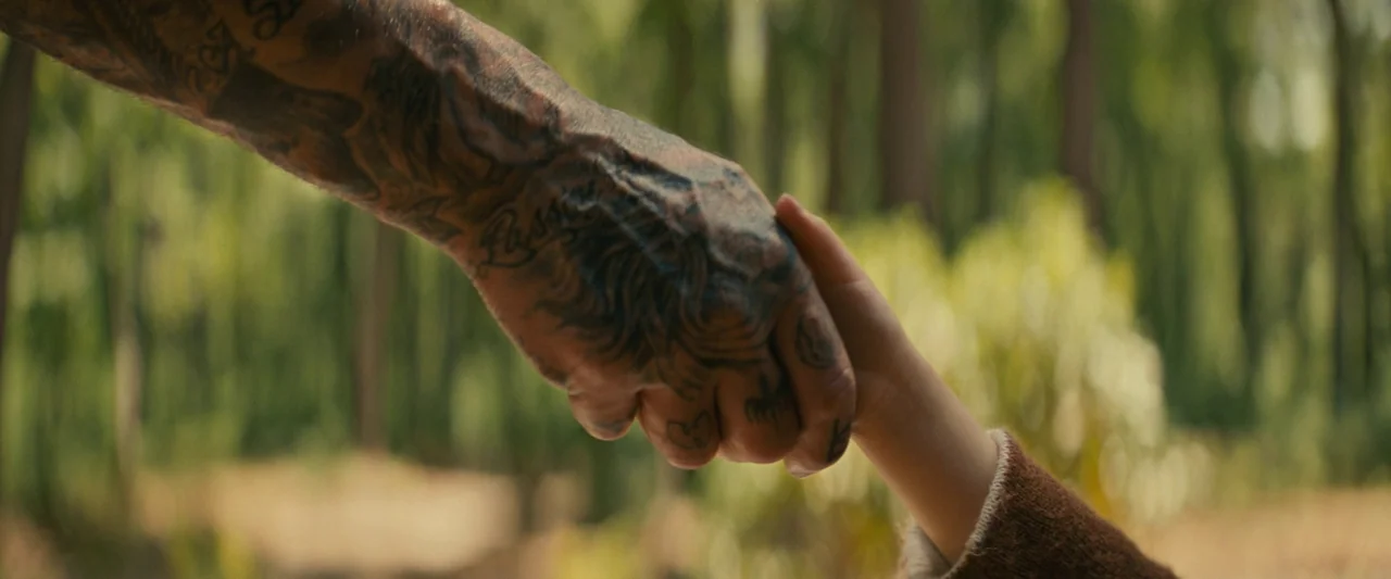Knock at the Cabin (2023) movie still: closeup — A close-up of a heavily tattooed adult hand holding a child's hand. The background consists of a…
