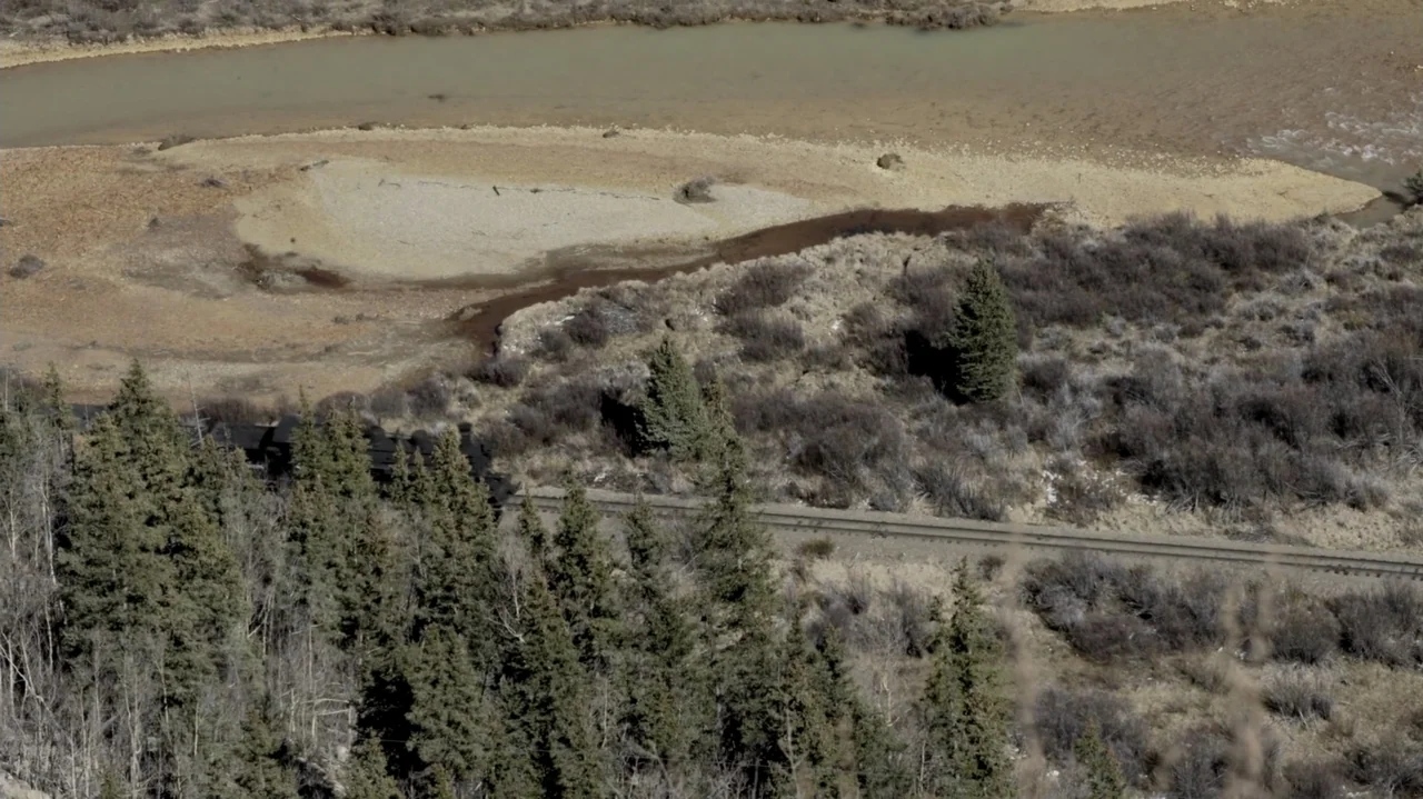 The Dalton Gang (2020) movie still: wide — A rural riverbank features sparse vegetation, coniferous trees, and active railroad tracks. A black steam…