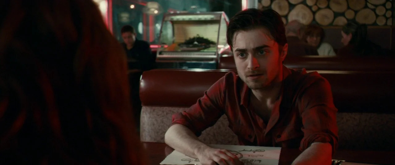 Horns (2013) movie still: medium — A man sits at a dark wooden booth inside a diner with wood-paneled walls. He focuses his gaze on an unseen person…