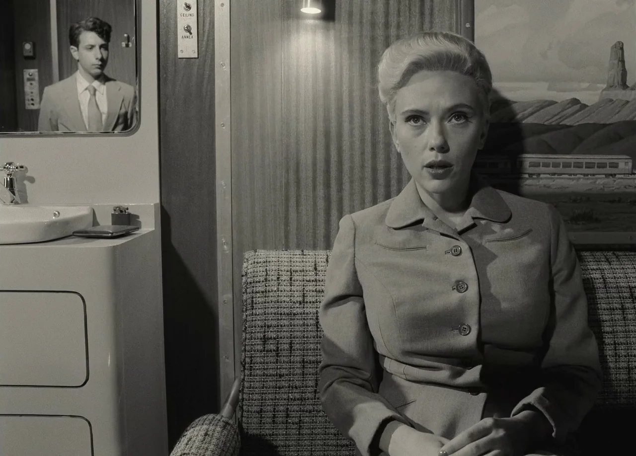 Asteroid City (2023) movie still: medium — A cramped train cabin features a small sink and a mirror reflecting a man. A woman sits on a patterned…