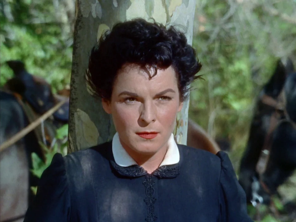 Johnny Guitar (1954) movie still: closeup — A woman leans against a thick tree trunk in a dense, green woodland setting. A black horse stands…