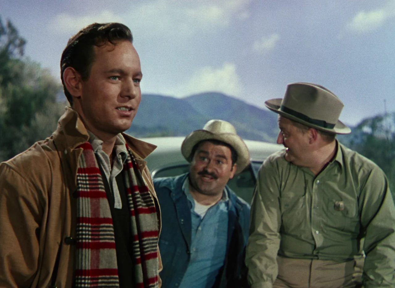 The War of the Worlds (1953) movie still: medium — A rural setting with distant rolling mountains visible behind a parked vintage automobile. Three…