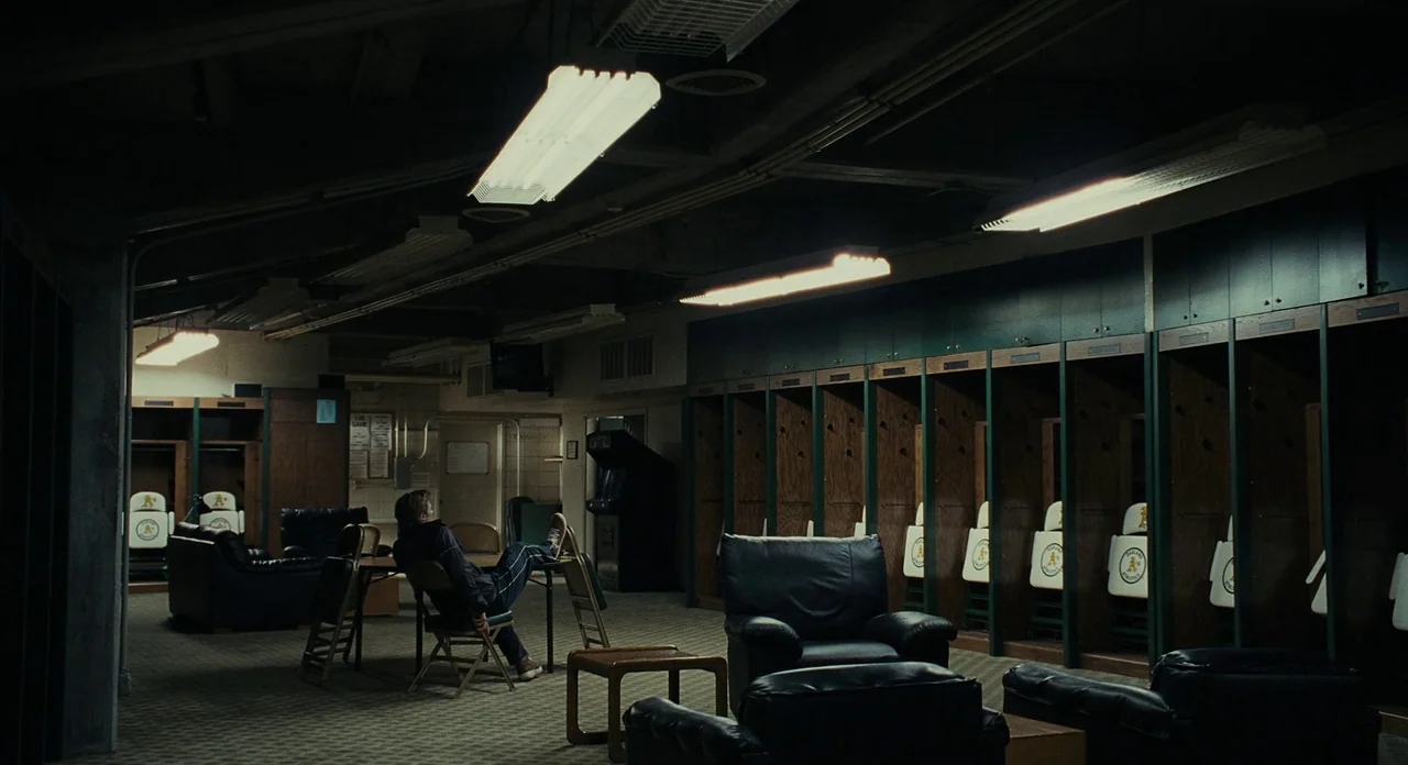 Moneyball (2011) movie still: wide — A dark, dimly lit professional sports team dressing room with rows of wooden lockers. A single man sits alone…