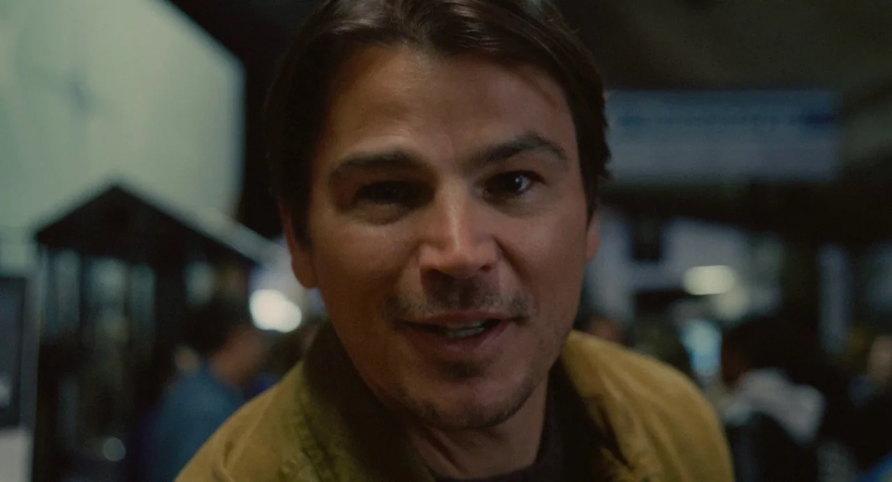 Trap (2024) movie still: closeup — A man stands in a bustling public transport hub with blurred background activity. He faces the camera directly…