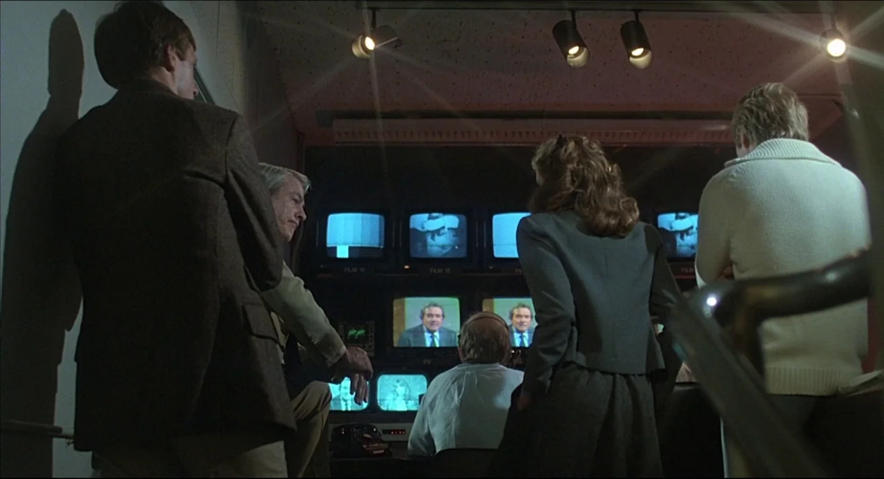 The Howling (1981) movie still: medium — A dimly lit television control room features walls of glowing video monitors. Several people stand in the…