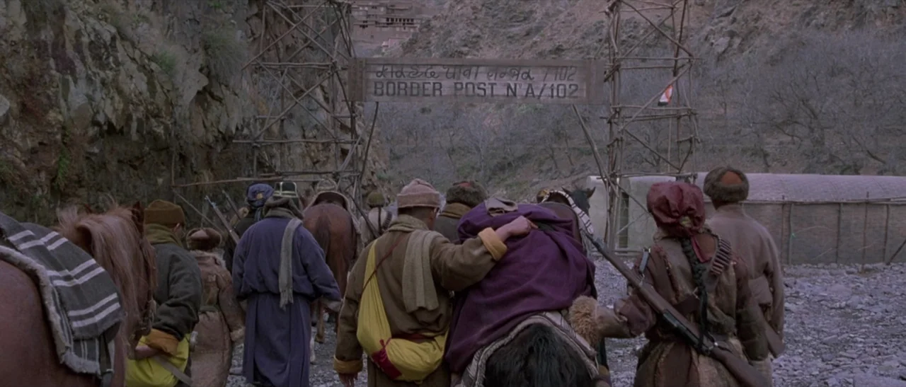Kundun (1997) movie still: wide — A rocky mountain pass features a rustic wooden sign marking a border post. A group of people walks away from the…