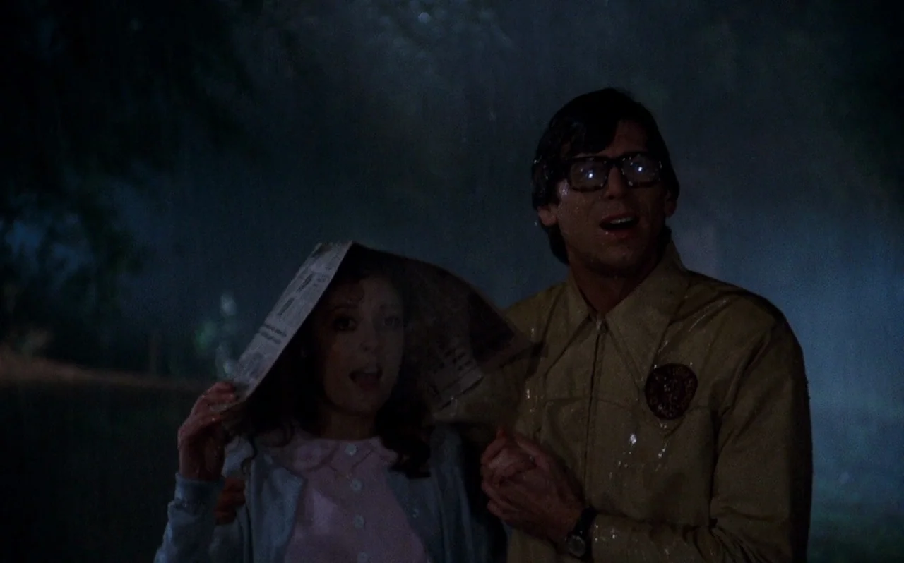 The Rocky Horror Picture Show (1975) movie still: medium — A dark, rainy outdoor area at night with foliage visible in the periphery. A man and a…