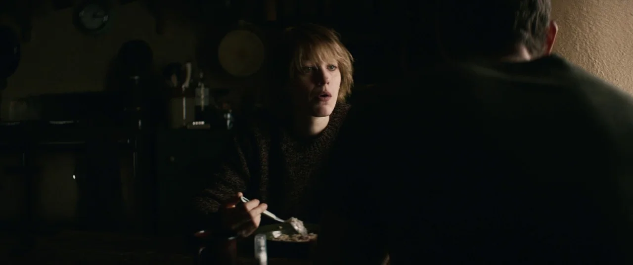 The Rhythm Section (2020) movie still: medium — A dimly lit room with wooden furniture and wall-mounted objects obscured in shadow. A woman sits at…
