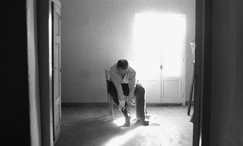 The Man From London (2007) movie still: fullbody — A sparsely furnished room with light walls and a wooden floor viewed through a doorway. A man…