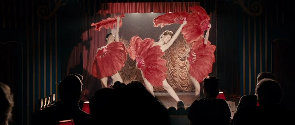 The Look of Love (2013) movie still: medium — A dark cabaret room features a small wooden stage with gold curtains and wall paneling. Three women…