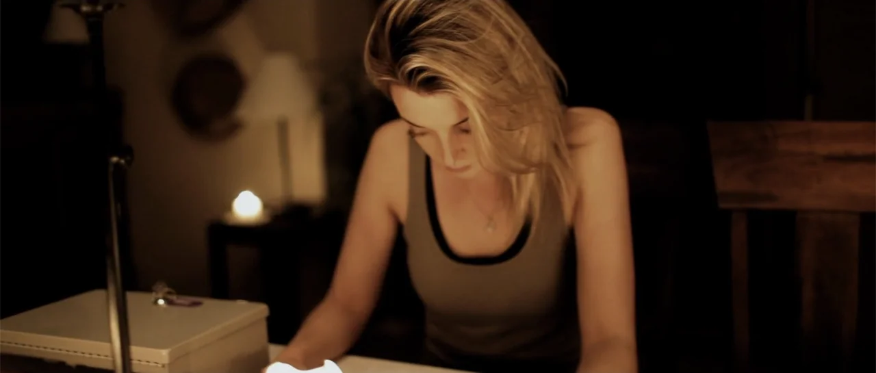 La coherencia del caos movie still: medium — A dimly lit room features a woman sitting at a table with a white surface. The woman with blonde hair…