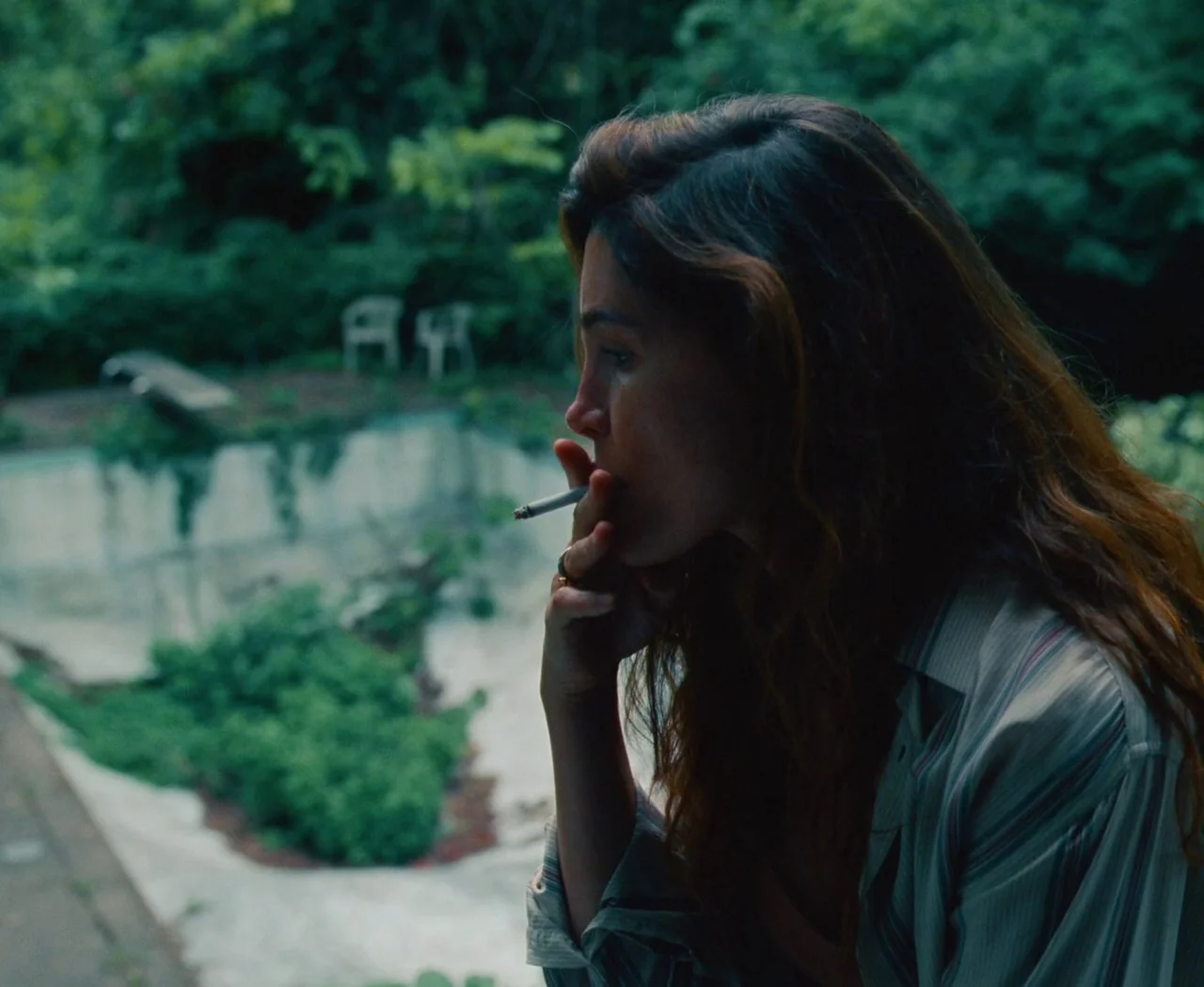 Monica (2022) movie still: medium — An overgrown, empty concrete pool sits in the background of a residential yard. A young woman in a striped shirt…