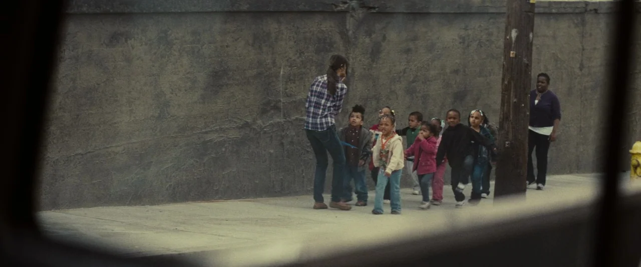 Out of the Furnace (2013) movie still: wide — A sidewalk sits beside a tall, weathered concrete wall near a wooden utility pole. A young woman walks…