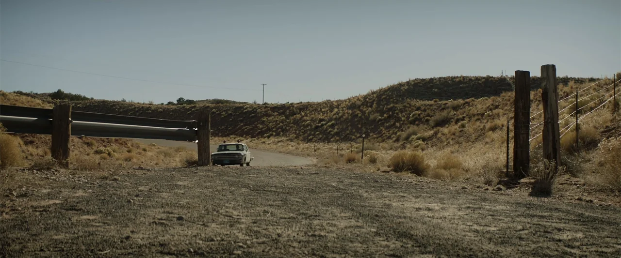 Cry Macho (2021) movie still: wide — A dry desert landscape features a winding paved road bordered by hills and fencing. A classic American sedan…