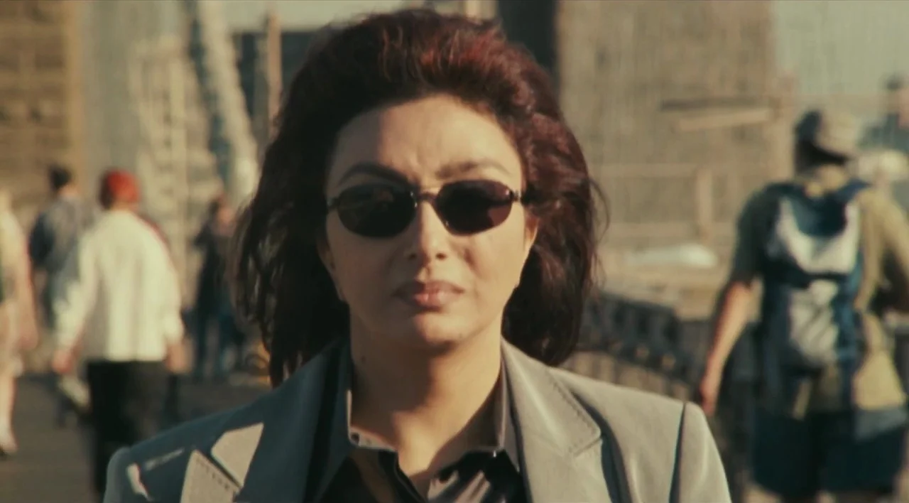 The Other Conquest (1999) movie still: closeup — A woman walks across a busy urban bridge filled with blurred pedestrian activity. The subject moves…