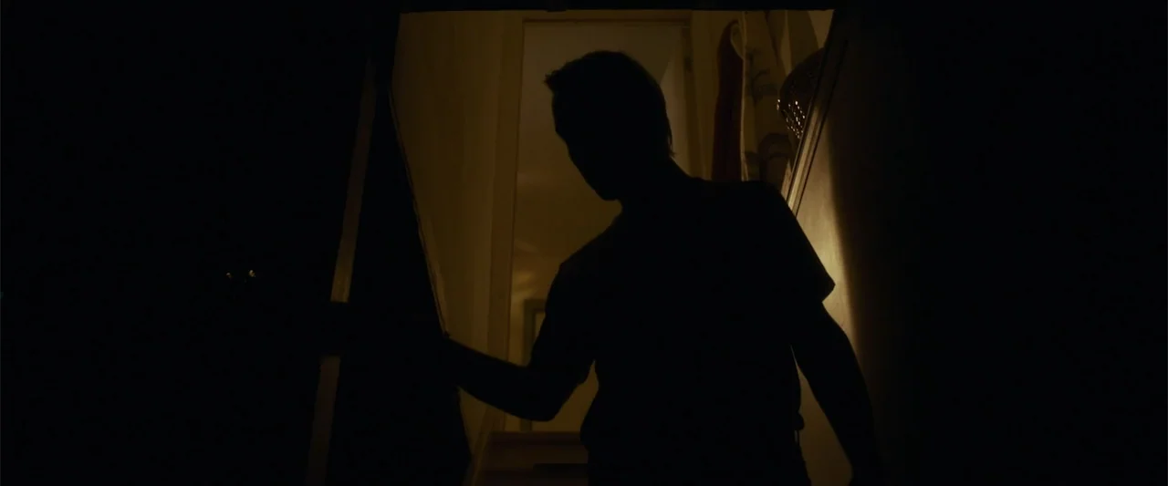 Jump, Darling (2022) movie still: medium — A dark, narrow indoor stairwell features a doorway leading into a brightly lit room. A person stands in…