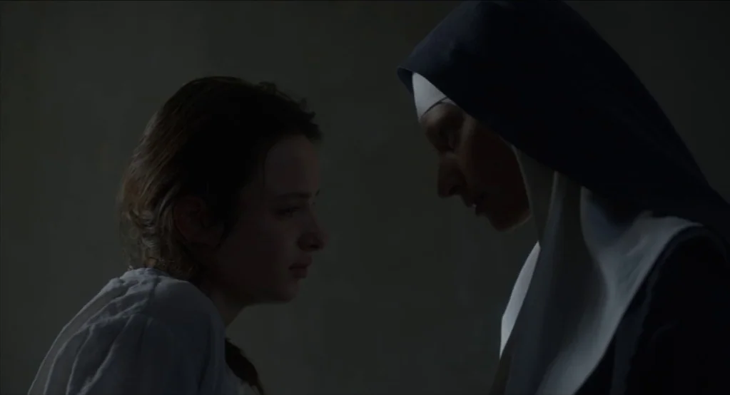The Innocents (2016) movie still: medium — A dimly lit, plain room with dark, indistinct walls surrounding the two figures. A young woman and a nun…