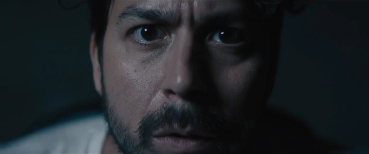 Stalker (2020) movie still: closeup — A dimly lit interior space with a dark, indistinct background. A man stares directly toward the camera with…