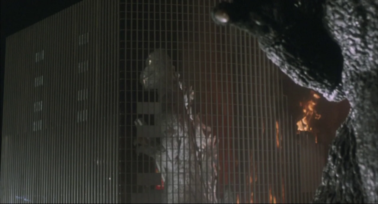 The Return of Godzilla (1984) movie still: wide — A gargantuan monster stands before a massive office building with a metallic grid facade. Flames…