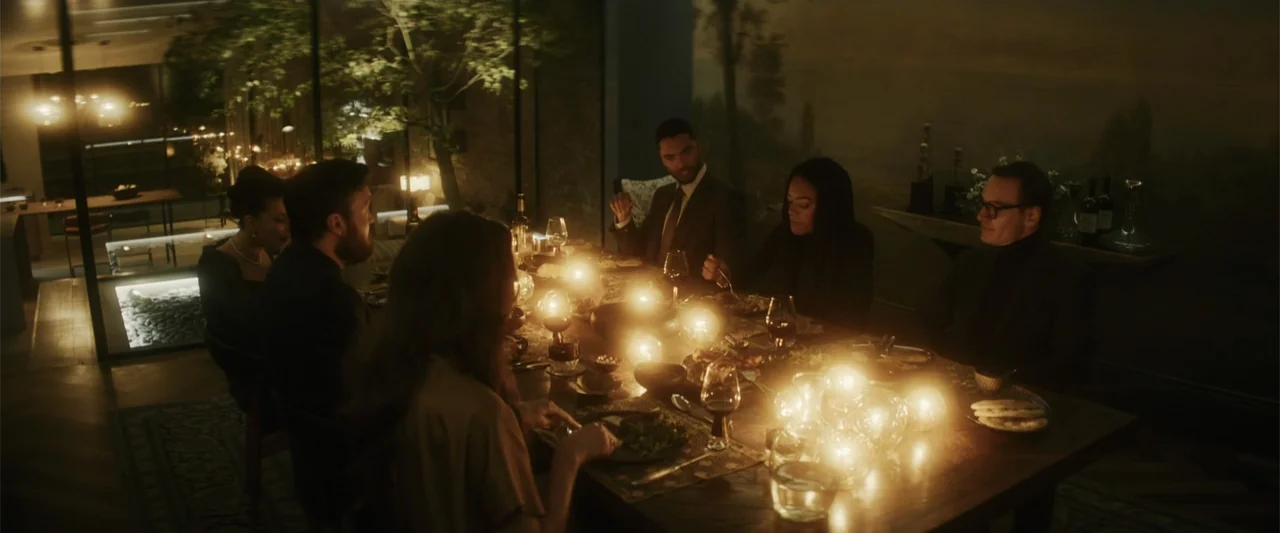 Black Bag (2025) movie still: wide — A dark dining room with a large wooden table illuminated by multiple spherical lamps. Six people are gathered…