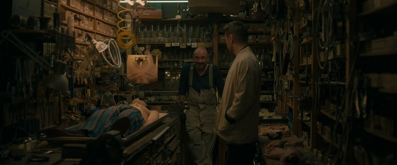 After Yang (2021) movie still: medium — A dark workshop is filled with shelves of hardware, tools, and mechanical components. An elderly man in…