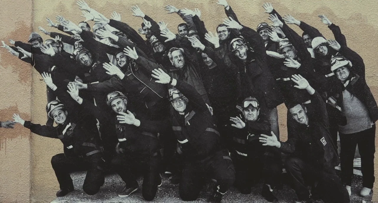 Faces Places (2017) movie still: wide — A group of people wearing safety helmets and uniforms pose against a textured wall. They raise their arms…