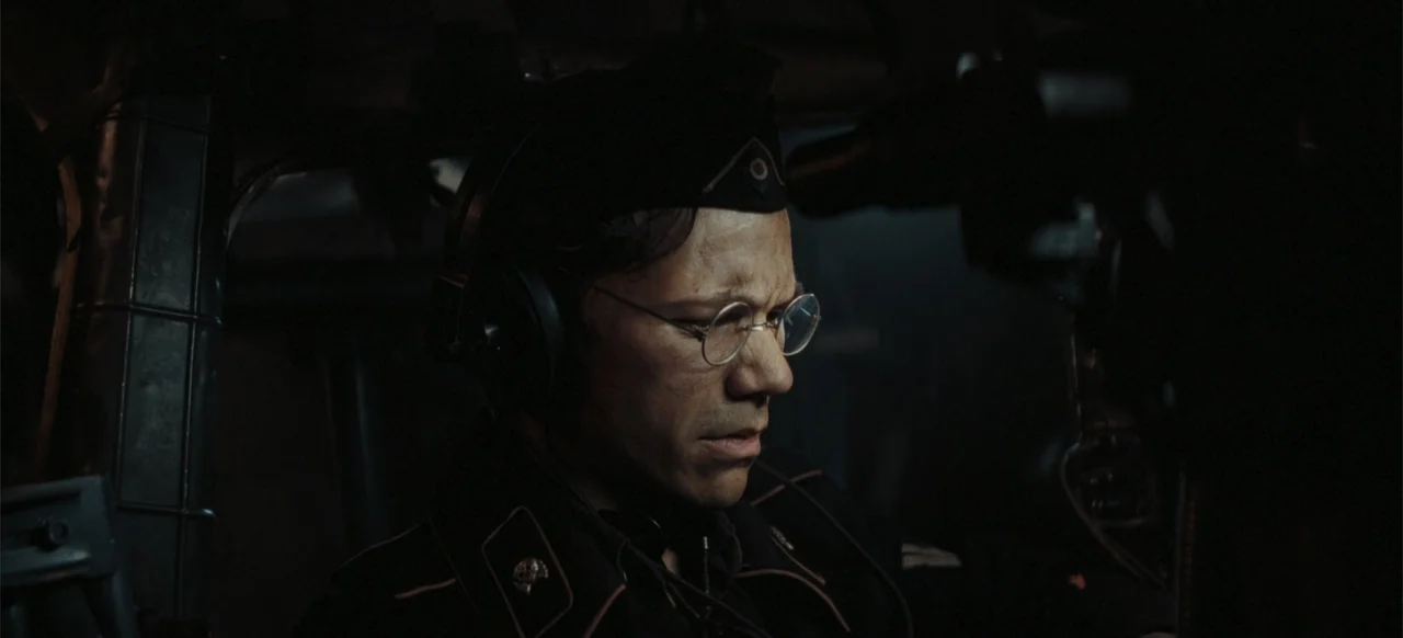 The Tank (2025) movie still: closeup — A dark, cramped military cockpit interior features mechanical controls and dim metallic surfaces. A man in a…